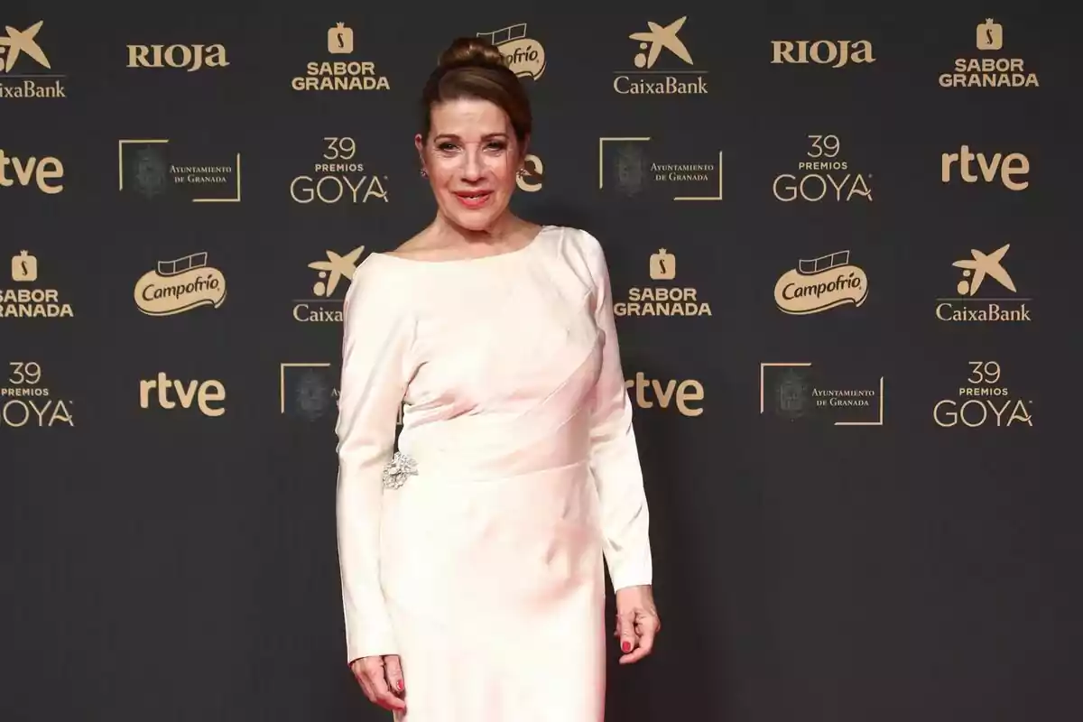 Una persona posando en la alfombra roja de los Premios Goya con un vestido elegante de color claro y un fondo con logotipos de patrocinadores. Una persona posando en la alfombra roja de los Premios Goya con un vestido elegante de color claro y un fondo con logotipos de patrocinadores.