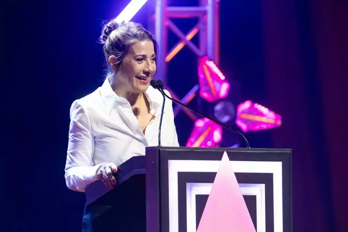 Una mujer con camisa blanca habla en un podio durante un evento con luces de colores en el fondo. Una mujer con camisa blanca habla en un podio durante un evento con luces de colores en el fondo.