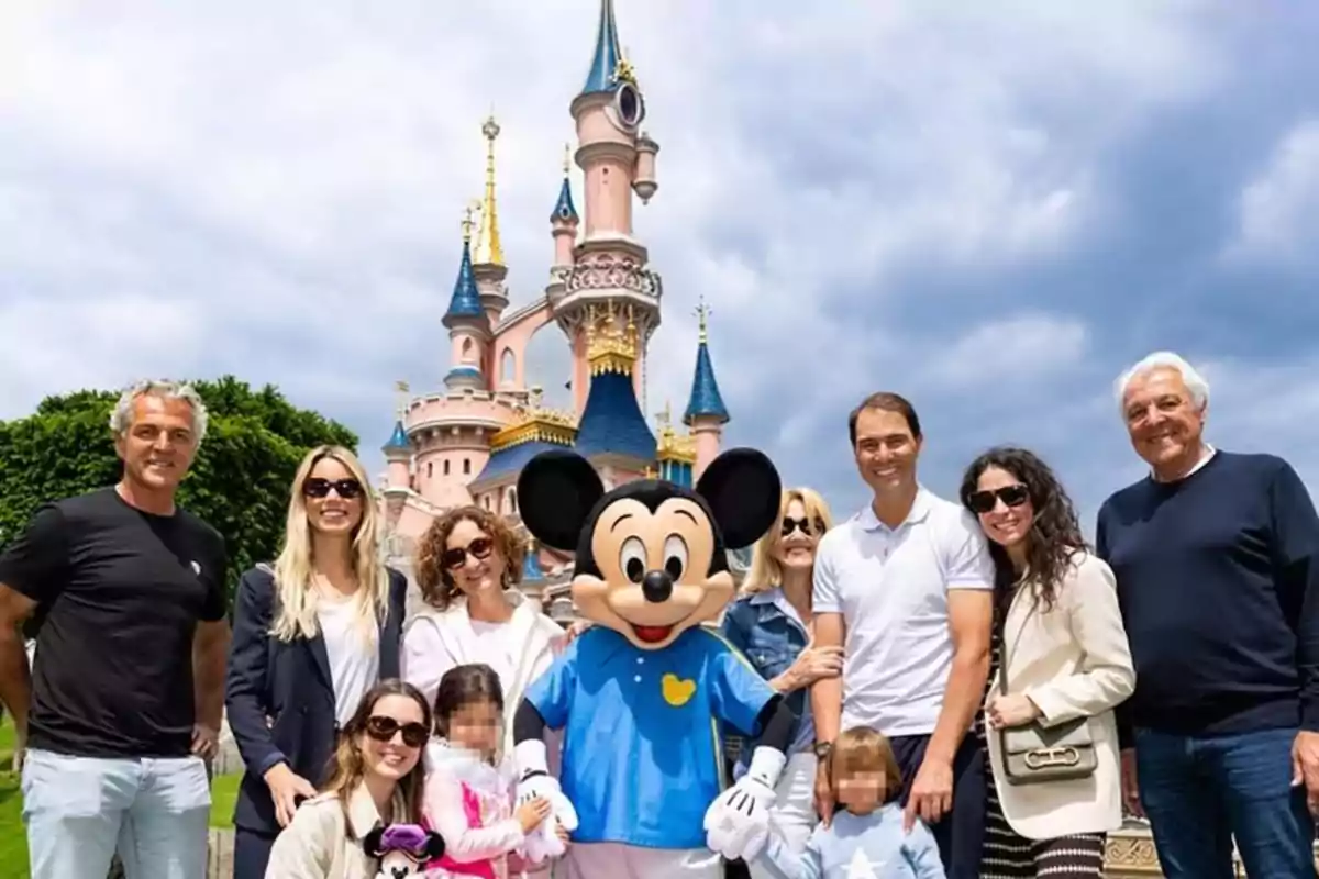 Un grup de persones somriu al costat d’un personatge disfressat de Mickey Mouse davant d’un castell en un parc temàtic de Disney sota un cel ennuvolat. Un grup de persones somriu al costat d’un personatge disfressat de Mickey Mouse davant d’un castell en un parc temàtic de Disney sota un cel ennuvolat.