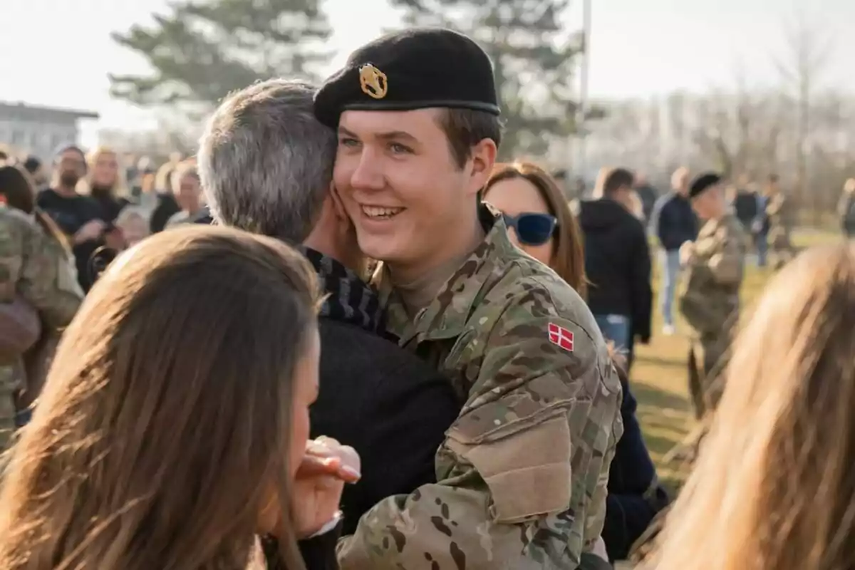 Un jove soldat amb uniforme militar somriu mentre abraça una persona en un esdeveniment a l'aire lliure amb més persones al voltant. Un jove soldat amb uniforme militar somriu mentre abraça una persona en un esdeveniment a l'aire lliure amb més persones al voltant.