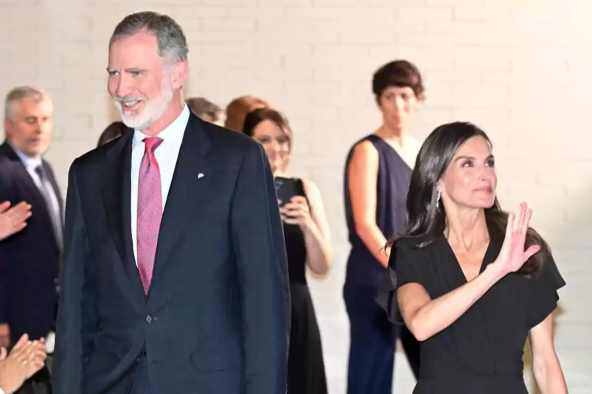 Un home amb vestit fosc i corbata vermella somriu mentre una dona vestida de negre saluda amb la mà, al fons hi ha diverses persones observant i una d’elles fa una foto amb el mòbil. Un home amb vestit fosc i corbata vermella somriu mentre una dona vestida de negre saluda amb la mà, al fons hi ha diverses persones observant i una d’elles fa una foto amb el mòbil.