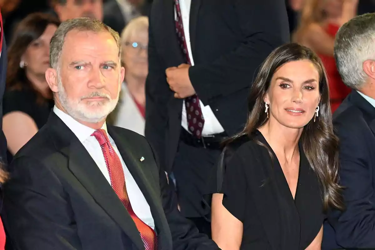 Un home amb barba i vestit fosc al costat d’una dona de cabell llarg i vestit negre asseguts en un esdeveniment formal envoltats d’altres persones Un home amb barba i vestit fosc al costat d’una dona de cabell llarg i vestit negre asseguts en un esdeveniment formal envoltats d’altres persones