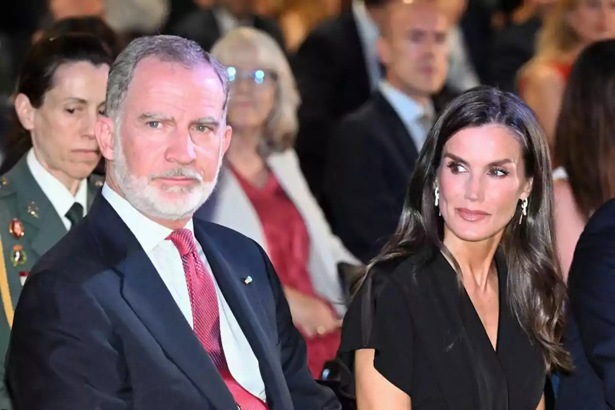 Un home amb vestit fosc i corbata vermella està assegut al costat d’una dona de cabells llargs i foscos vestida de negre en un esdeveniment formal envoltats d’altres persones. Un home amb vestit fosc i corbata vermella està assegut al costat d’una dona de cabells llargs i foscos vestida de negre en un esdeveniment formal envoltats d’altres persones.