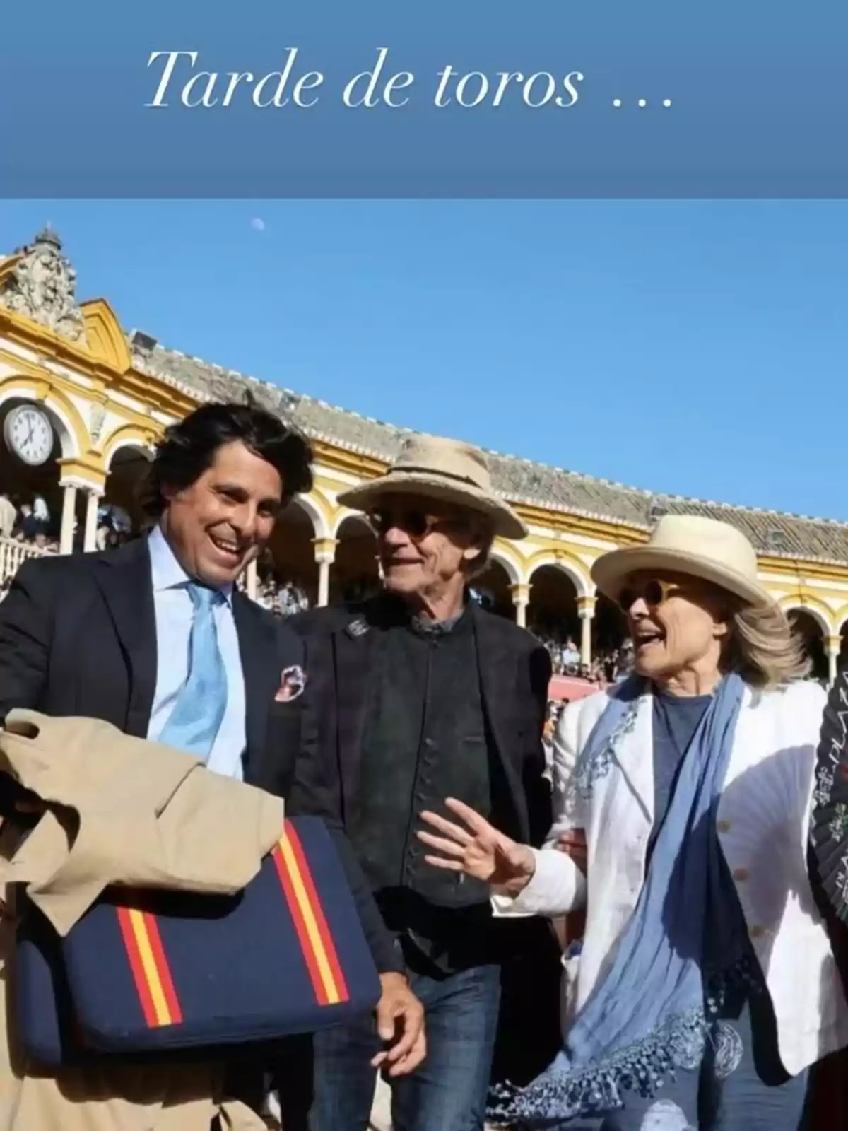 Tres persones somrients conversen en una plaça de toros, amb un edifici de fons i un cel clar. Tres persones somrients conversen en una plaça de toros, amb un edifici de fons i un cel clar.