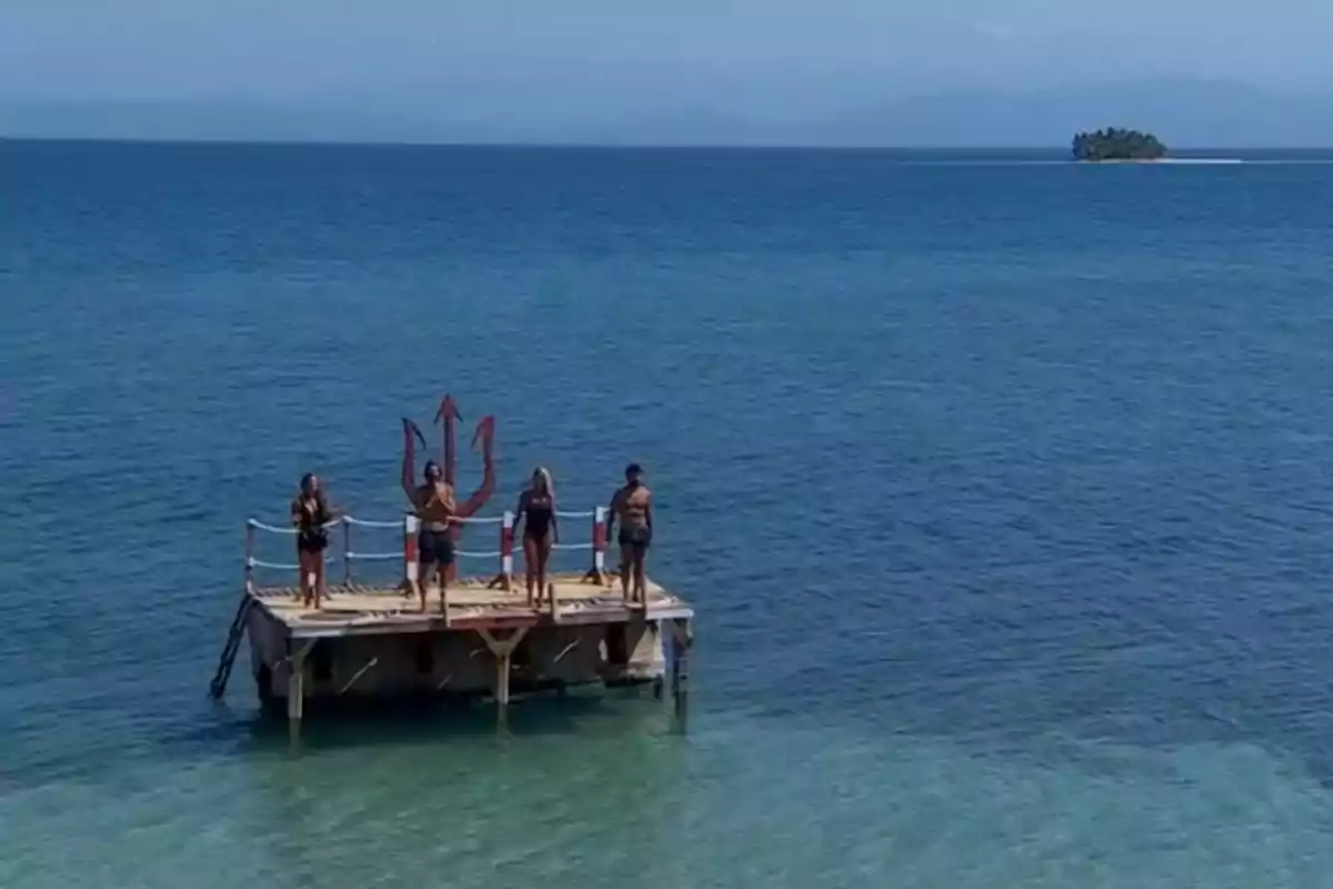Un grup de persones està dret sobre una plataforma al mar amb un trident gran darrere d'ells i una petita illa es veu a la llunyania. Un grup de persones està dret sobre una plataforma al mar amb un trident gran darrere d'ells i una petita illa es veu a la llunyania.