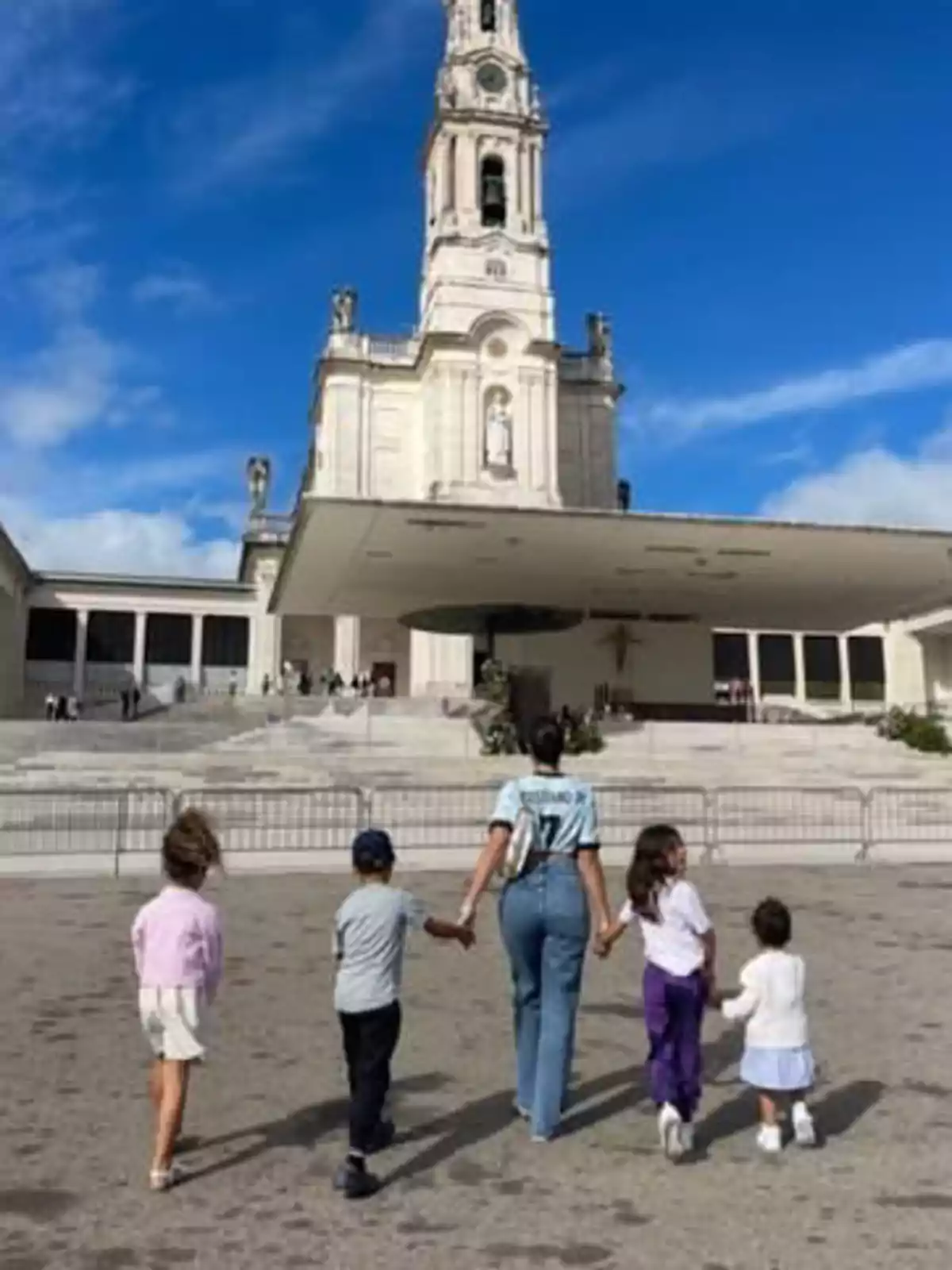 Una dona i quatre nens caminen de la mà cap a un edifici religiós amb una torre alta sota un cel blau. Una dona i quatre nens caminen de la mà cap a un edifici religiós amb una torre alta sota un cel blau.