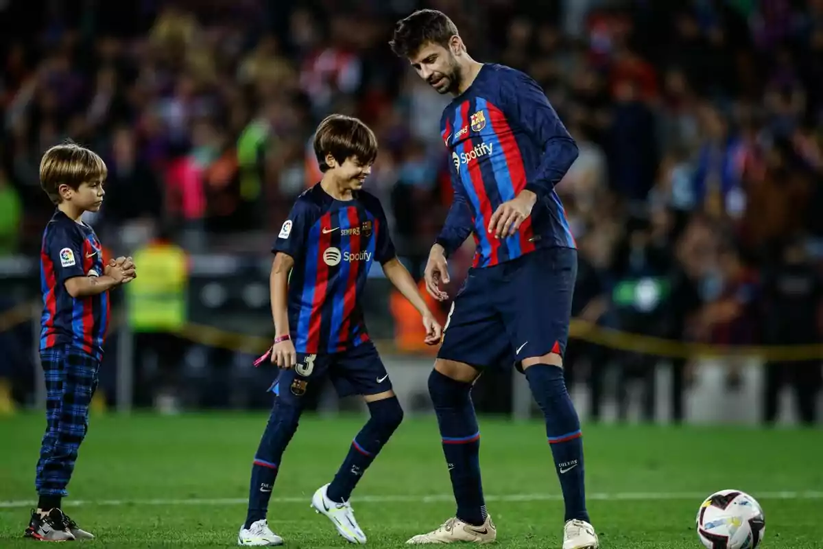 A player from FC Barcelona is on the field with two children, all wearing the team uniform, while interacting and smiling. A player from FC Barcelona is on the field with two children, all wearing the team uniform, while interacting and smiling.