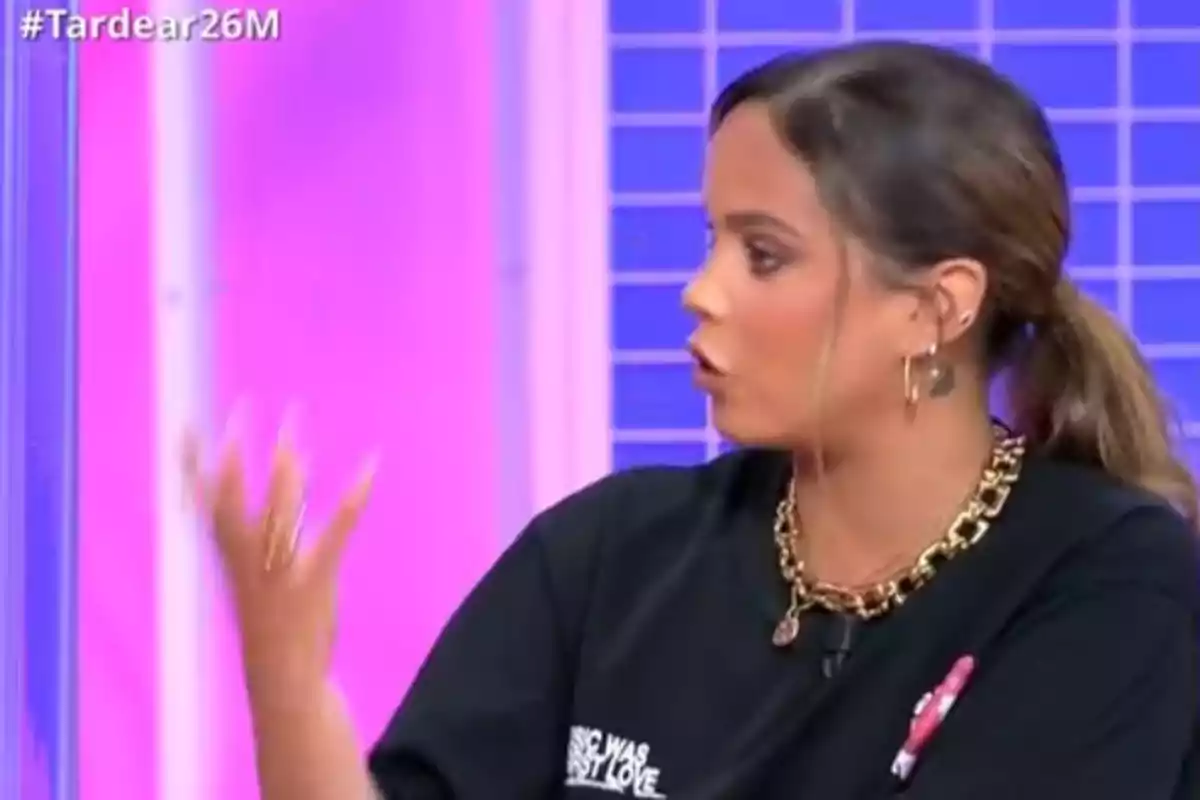 Dona jove amb cua i samarreta negra parlant i gesticulant en un plató de televisió amb fons rosa i blau Dona jove amb cua i samarreta negra parlant i gesticulant en un plató de televisió amb fons rosa i blau