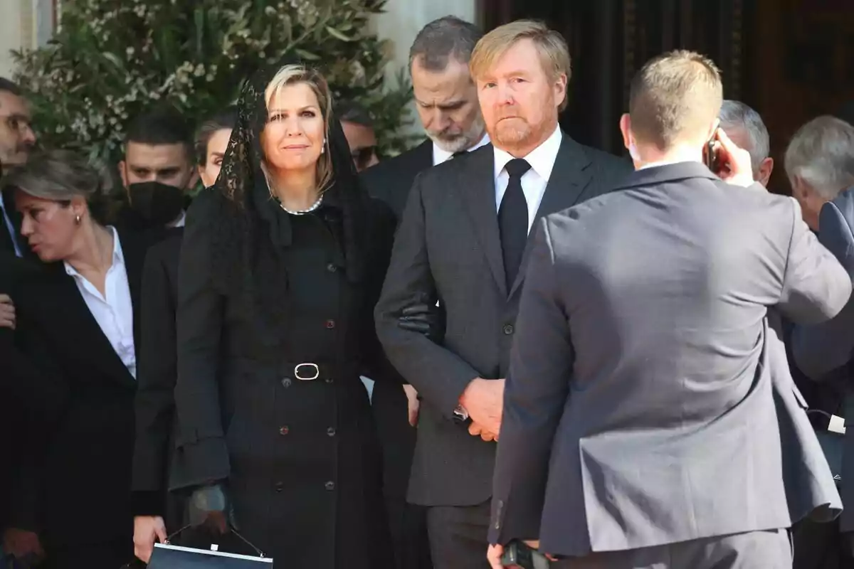 Un grup de persones vestides de manera formal es troba reunit a l'aire lliure, algunes d'elles llueixen vestits foscos i una dona porta un vel negre sobre el cap. Un grup de persones vestides de manera formal es troba reunit a l'aire lliure, algunes d'elles llueixen vestits foscos i una dona porta un vel negre sobre el cap.