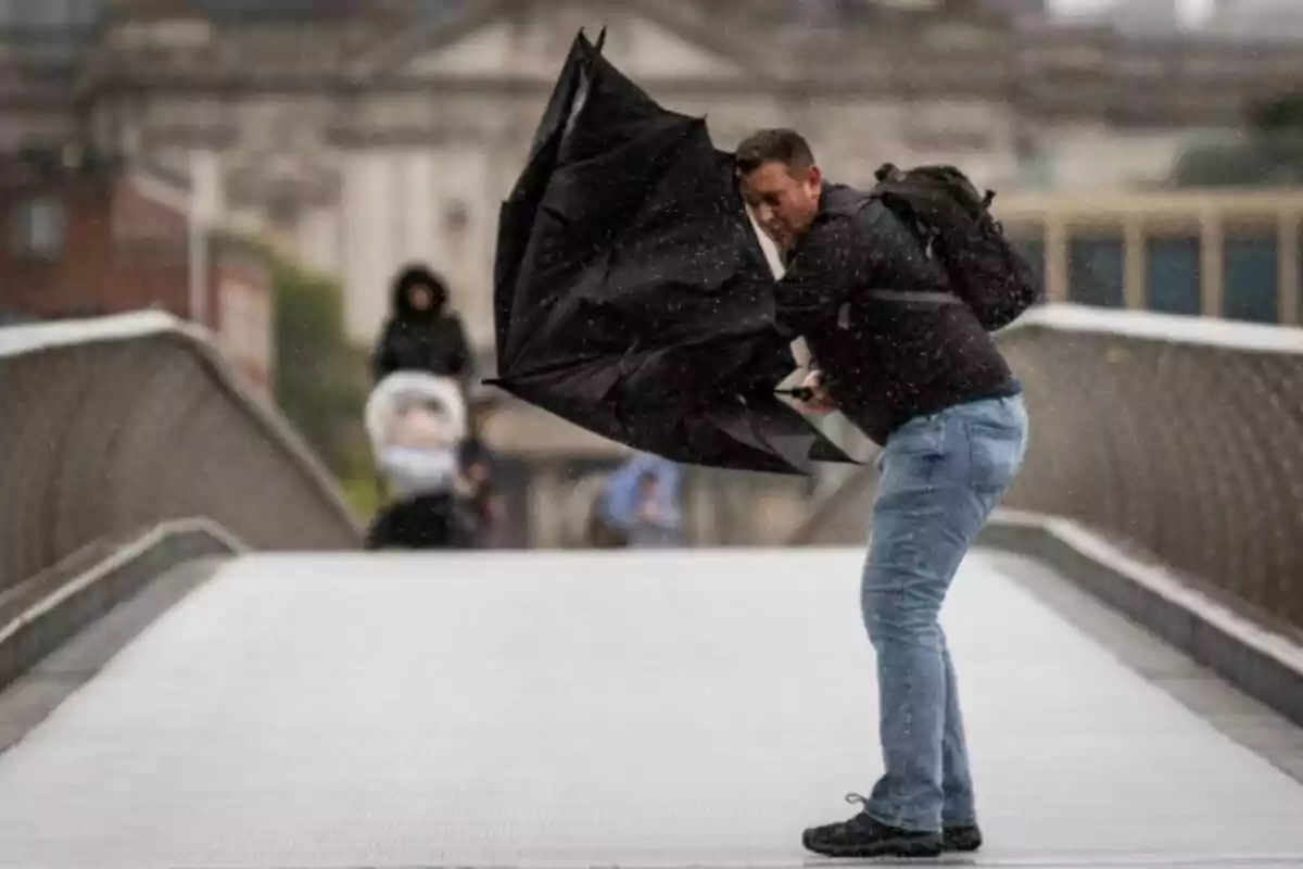 Hombre luchando contra el viento con un paraguas invertido en un puente durante un día lluvioso.