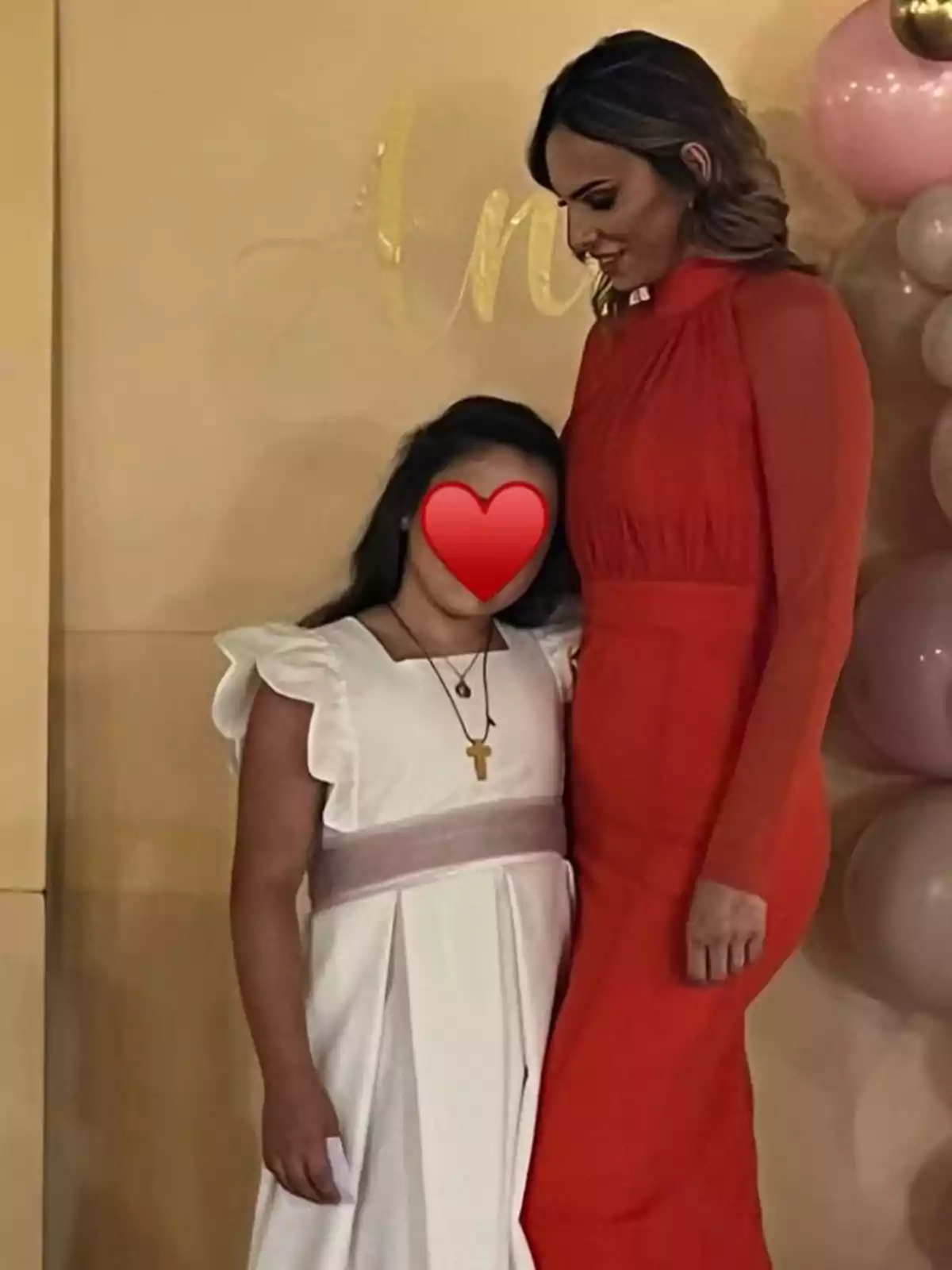 A girl in a white dress and a woman in a red dress pose together at a special event; the girl is wearing a cross necklace and her face is covered with a red heart. In the background, there are balloons and decorations in pastel colors. A girl in a white dress and a woman in a red dress pose together at a special event; the girl is wearing a cross necklace and her face is covered with a red heart. In the background, there are balloons and decorations in pastel colors.