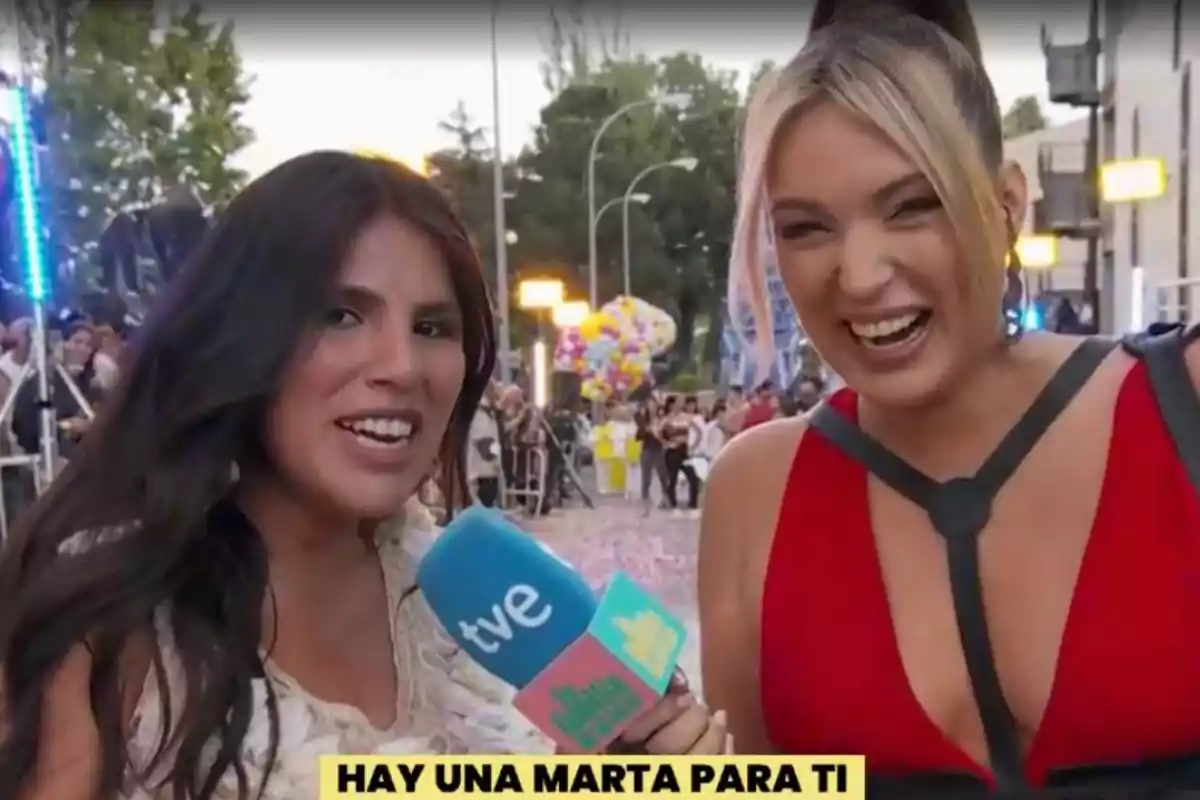 Dues dones somriuen mentre una d'elles sosté un micròfon de TVE en un esdeveniment a l'aire lliure amb globus i persones de fons. Dues dones somriuen mentre una d'elles sosté un micròfon de TVE en un esdeveniment a l'aire lliure amb globus i persones de fons.
