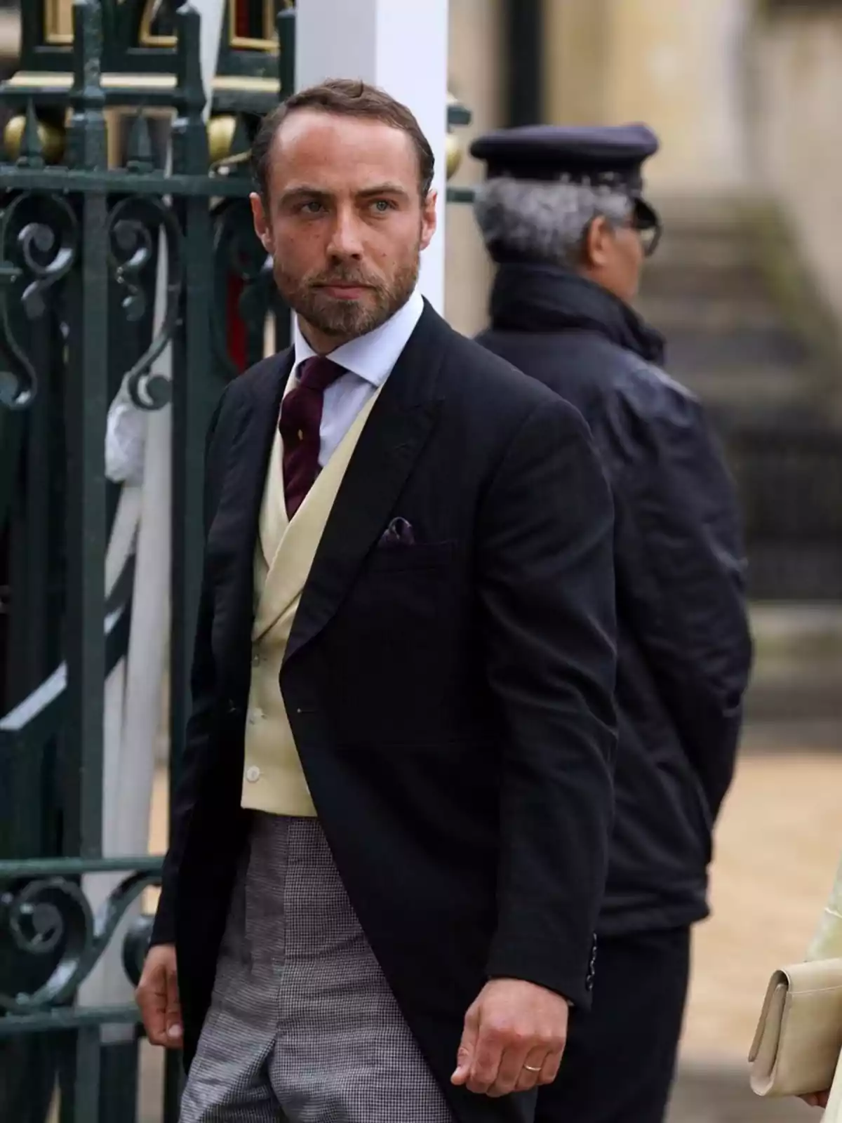 Un home amb barba i cabell curt, vestit amb un vestit formal de jaqueta negra, armilla beix, corbata vermella i pantalons a quadres, està dreta davant d'una reixa verda, mentre una persona amb uniforme fosc i gorra està d'esquena al fons . Un home amb barba i cabell curt, vestit amb un vestit formal de jaqueta negra, armilla beix, corbata vermella i pantalons a quadres, està dreta davant d'una reixa verda, mentre una persona amb uniforme fosc i gorra està d'esquena al fons .