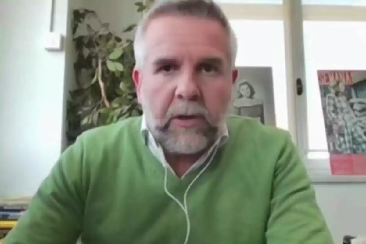 Un home amb barba i jersei verd apareix en una videotrucada, amb un fons que inclou una planta i cartells a la paret. Un home amb barba i jersei verd apareix en una videotrucada, amb un fons que inclou una planta i cartells a la paret.