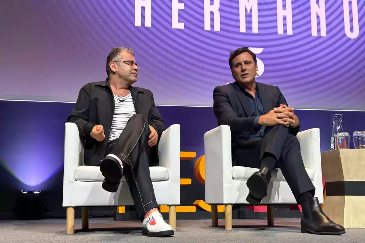 Dos homes asseguts en butaques blanques en un escenari, un amb jaqueta negra i samarreta de ratlles, l'altre amb vestit fosc, amb un fons que té lletres grans. Dos homes asseguts en butaques blanques en un escenari, un amb jaqueta negra i samarreta de ratlles, l'altre amb vestit fosc, amb un fons que té lletres grans.