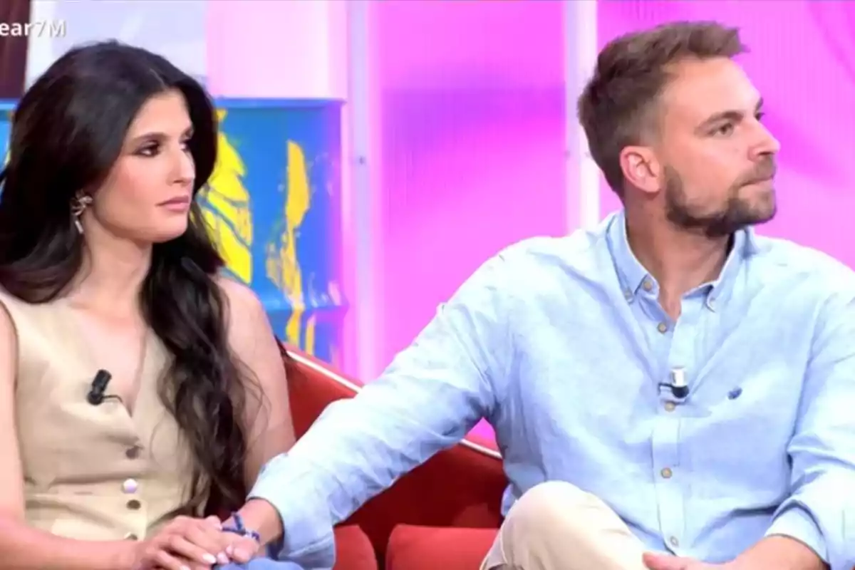 Una parella asseguda en un sofà vermell en un programa de televisió, tots dos miren cap a la dreta mentre es donen la mà. Una parella asseguda en un sofà vermell en un programa de televisió, tots dos miren cap a la dreta mentre es donen la mà.