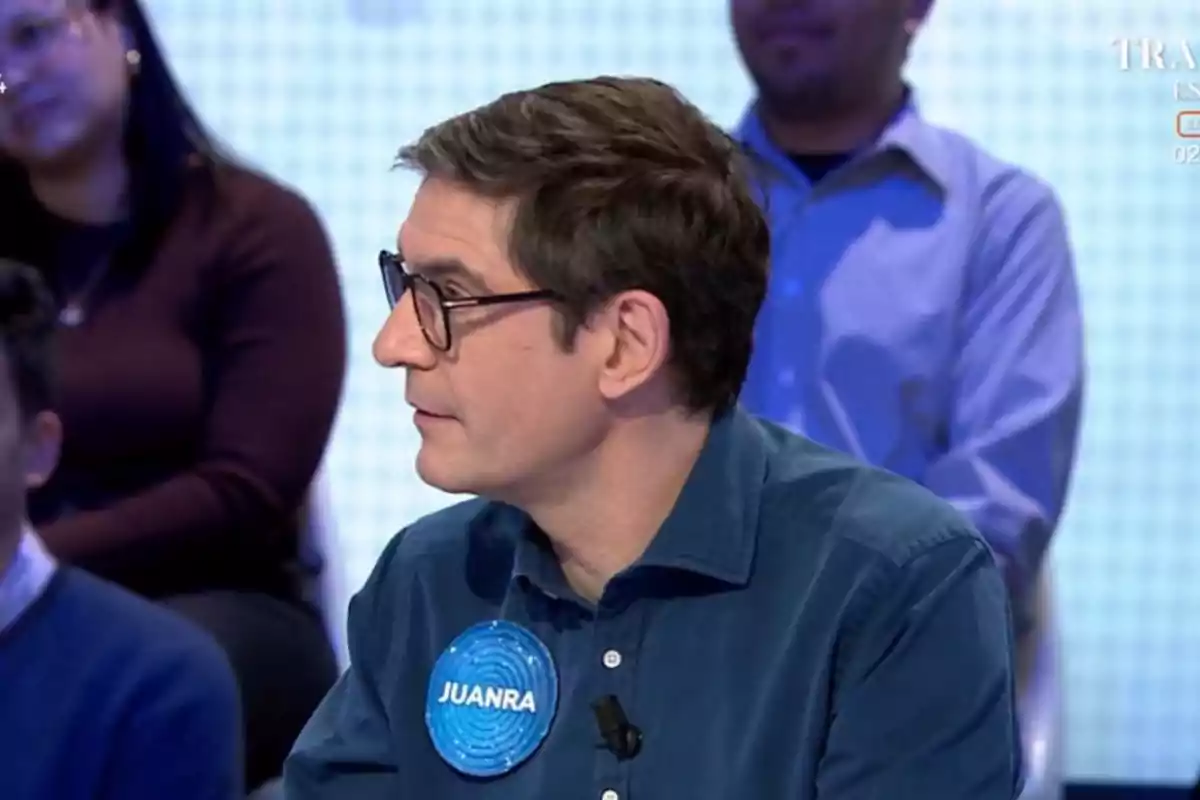 Un home amb ulleres i camisa blava participa en un programa de televisió, amb persones assegudes al fons. Un home amb ulleres i camisa blava participa en un programa de televisió, amb persones assegudes al fons.
