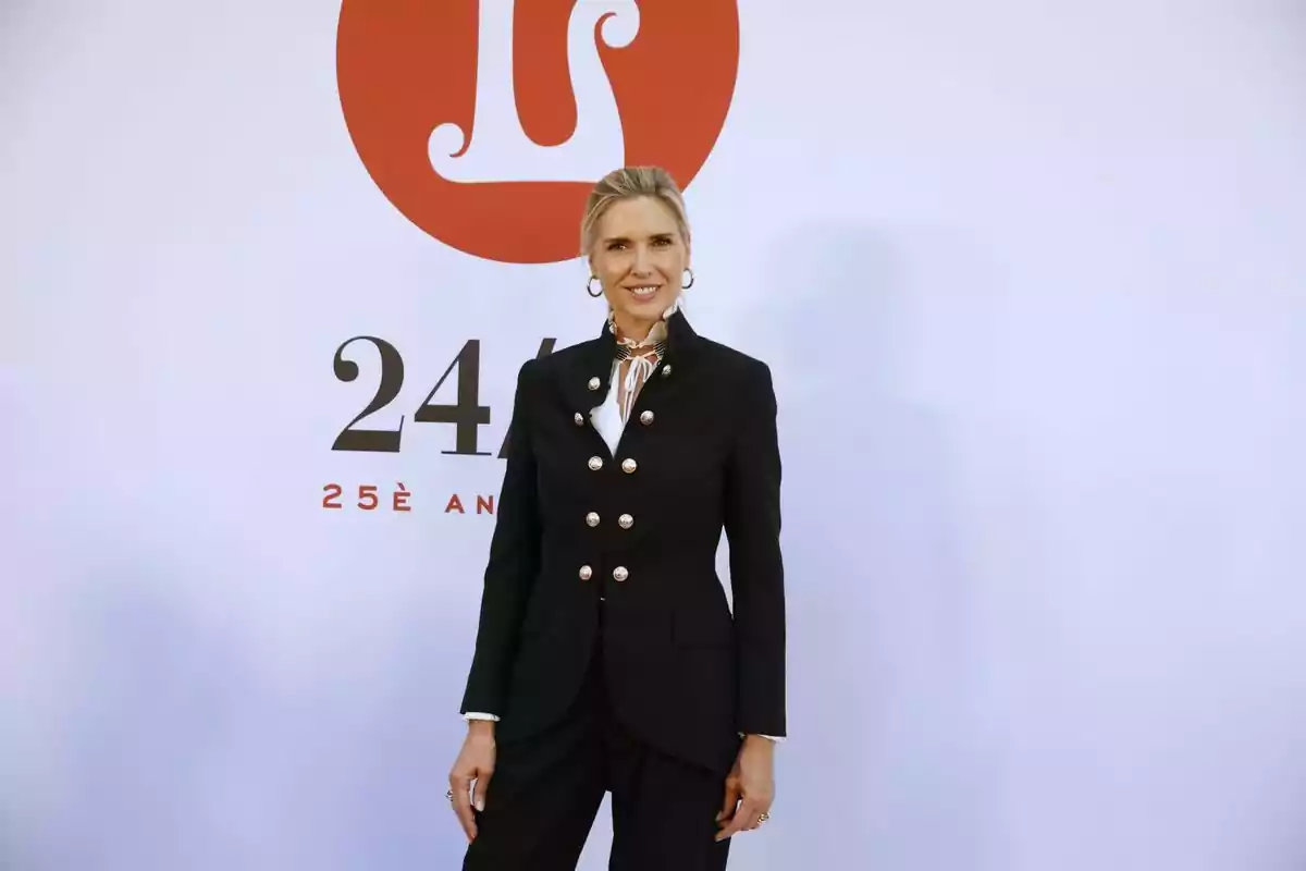 Una persona con traje negro posando frente a un fondo blanco con un logotipo rojo y números. Una persona con traje negro posando frente a un fondo blanco con un logotipo rojo y números.
