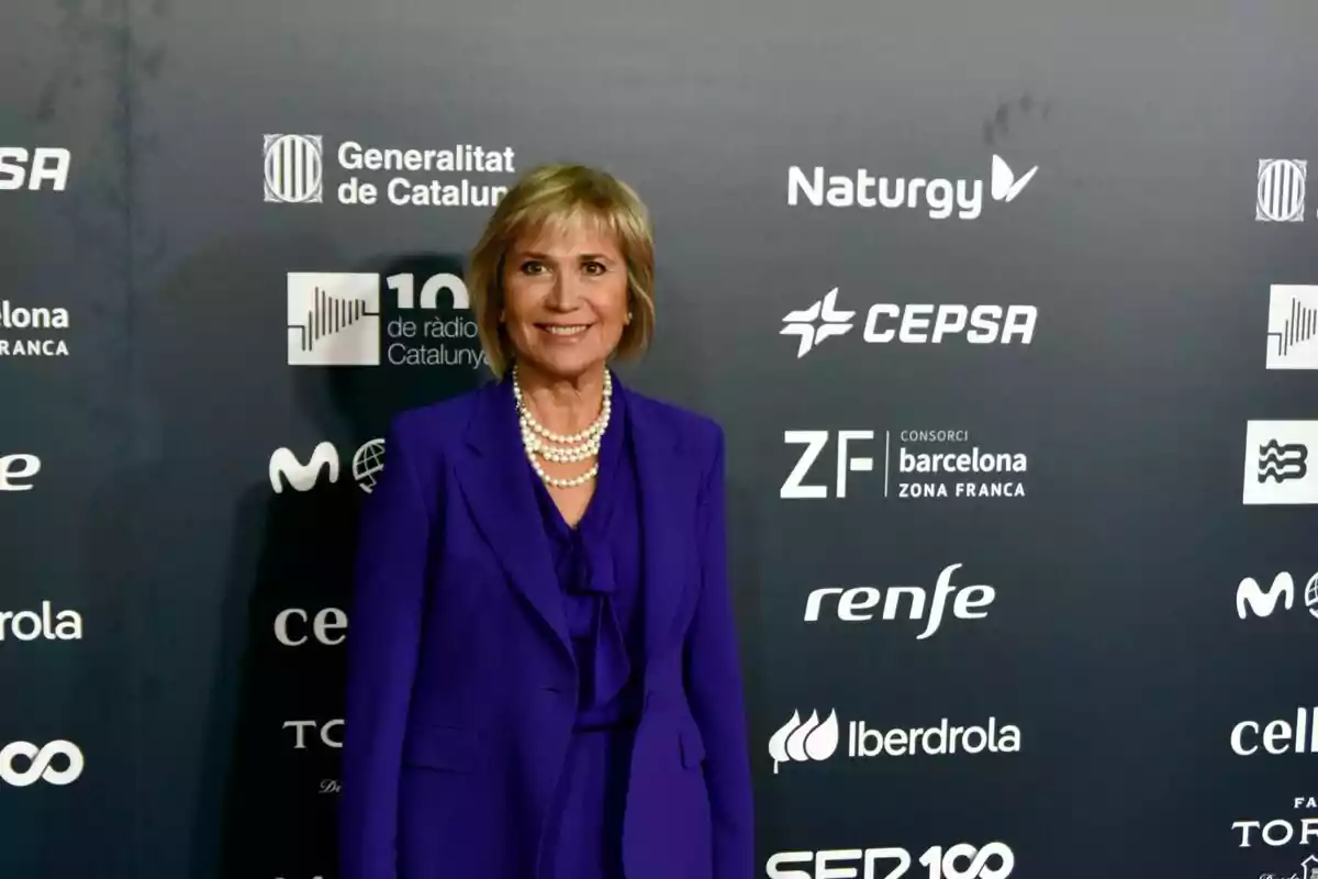 Una persona amb un vestit morat i un collaret de perles posa davant d'un fons amb logotips de diverses empreses i organitzacions. Una persona amb un vestit morat i un collaret de perles posa davant d'un fons amb logotips de diverses empreses i organitzacions.