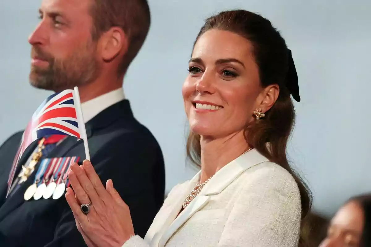 A smiling woman holds a small United Kingdom flag while standing next to a man with medals on his suit. A smiling woman holds a small United Kingdom flag while standing next to a man with medals on his suit.