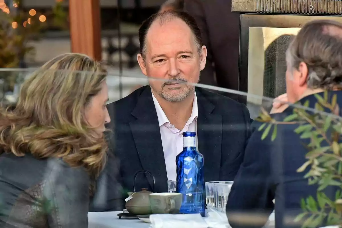 Three people are sitting at an outdoor restaurant table, talking, with a blue water bottle and cups on the table. Three people are sitting at an outdoor restaurant table, talking, with a blue water bottle and cups on the table.