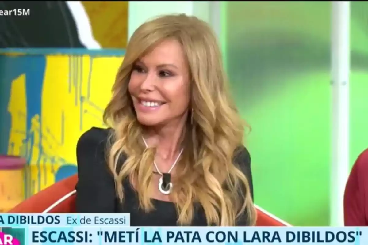 Una dona somrient amb cabell ros i ondulat està asseguda en un programa de televisió, amb un fons acolorit i un text a la part inferior de la imatge. Una dona somrient amb cabell ros i ondulat està asseguda en un programa de televisió, amb un fons acolorit i un text a la part inferior de la imatge.