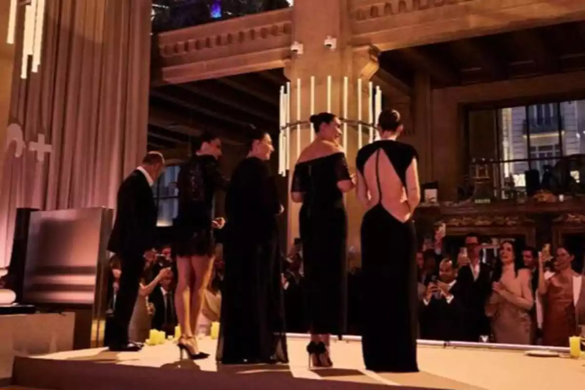 Un grup de persones elegantment vestides en un esdeveniment social en un saló amb il·luminació càlida i públic observant. Un grup de persones elegantment vestides en un esdeveniment social en un saló amb il·luminació càlida i públic observant.