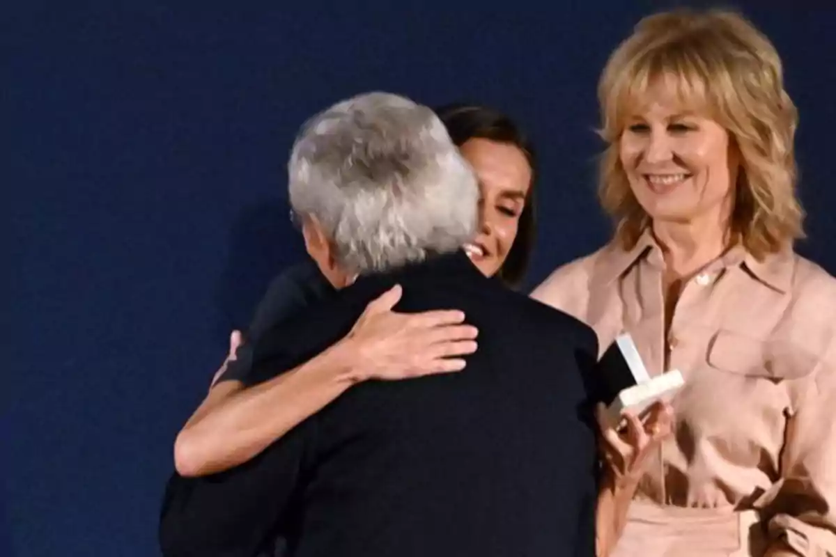 Una persona abraçant un home gran mentre una altra dona somriu sostenint una caixa petita. Una persona abraçant un home gran mentre una altra dona somriu sostenint una caixa petita.