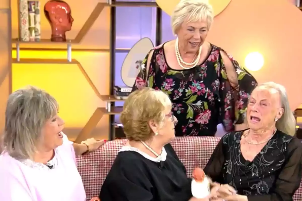 Un grup de dones grans està assegut en un sofà en un ambient d'estudi de televisió, una d'elles sembla emocionada mentre les altres l'observen i conversen. Un grup de dones grans està assegut en un sofà en un ambient d'estudi de televisió, una d'elles sembla emocionada mentre les altres l'observen i conversen.