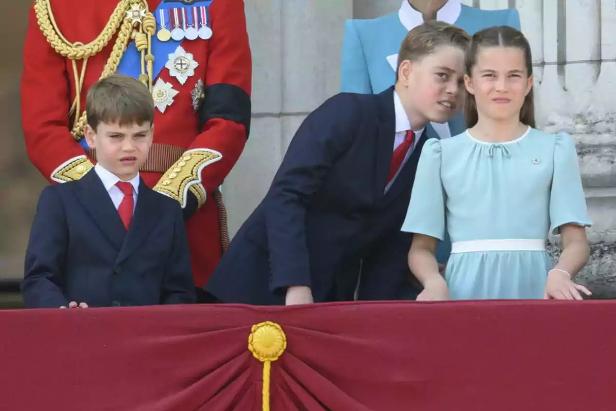 Three children dressed formally watch an event from a balcony decorated with red fabric and gold details, while elegantly dressed adults stand behind them. Three children dressed formally watch an event from a balcony decorated with red fabric and gold details, while elegantly dressed adults stand behind them.