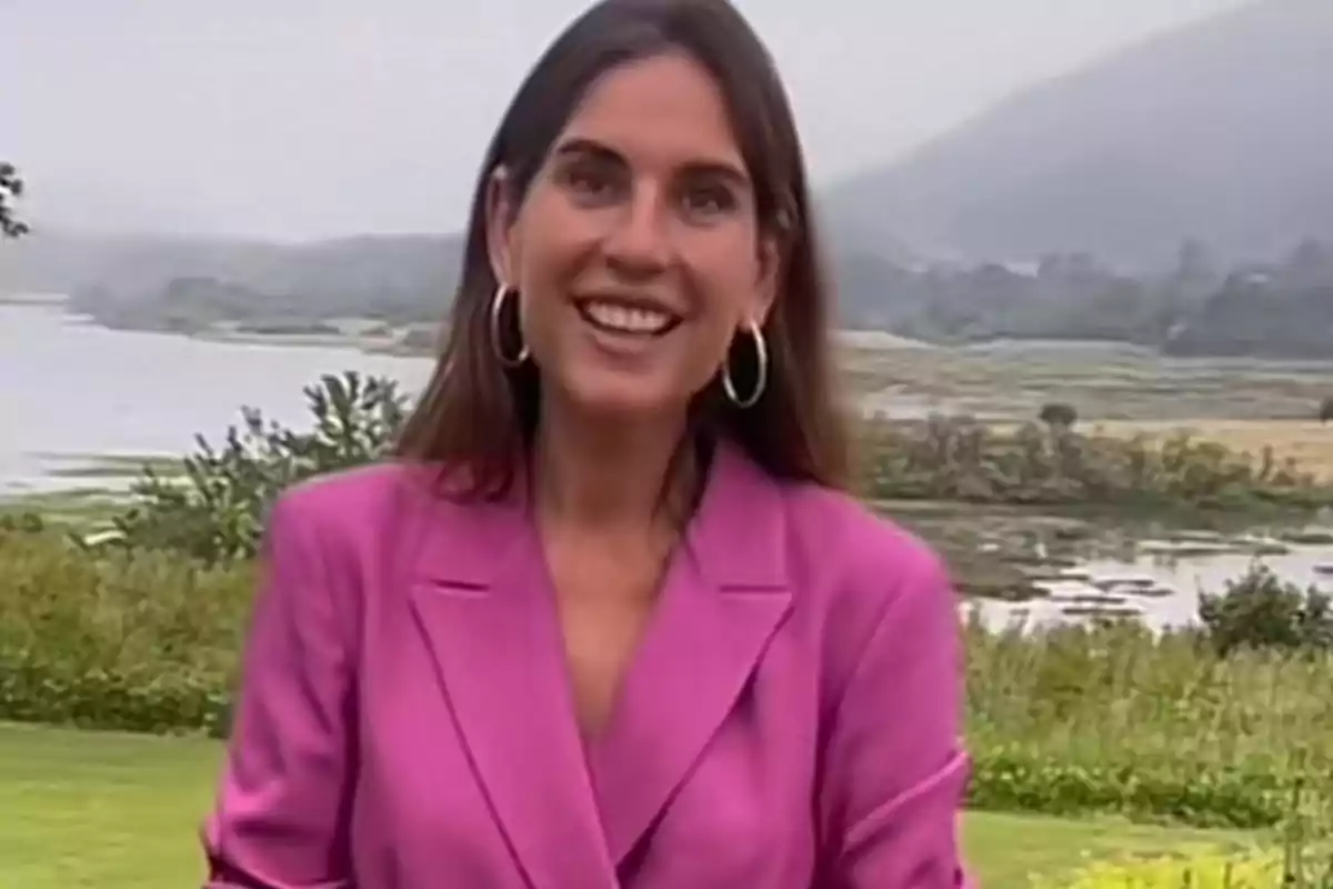 Dona somrient amb jaqueta rosa i grans arracades d’aro posant a l’aire lliure davant d’un paisatge de llac i muntanyes Dona somrient amb jaqueta rosa i grans arracades d’aro posant a l’aire lliure davant d’un paisatge de llac i muntanyes