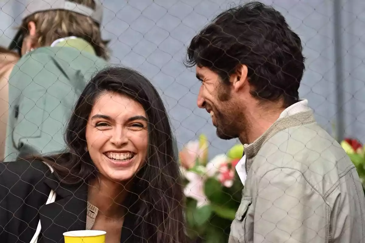 Una dona somrient sosté un got groc mentre conversa amb un home, tots dos estan darrere d'una reixa i semblen estar gaudint d'un esdeveniment a l'aire lliure. Una dona somrient sosté un got groc mentre conversa amb un home, tots dos estan darrere d'una reixa i semblen estar gaudint d'un esdeveniment a l'aire lliure.