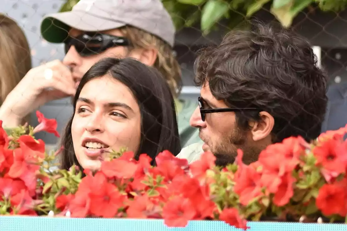 Un grup de persones està assegut darrere d'una fila de flors vermelles, conversant i gaudint d'un esdeveniment a l'aire lliure. Un grup de persones està assegut darrere d'una fila de flors vermelles, conversant i gaudint d'un esdeveniment a l'aire lliure.