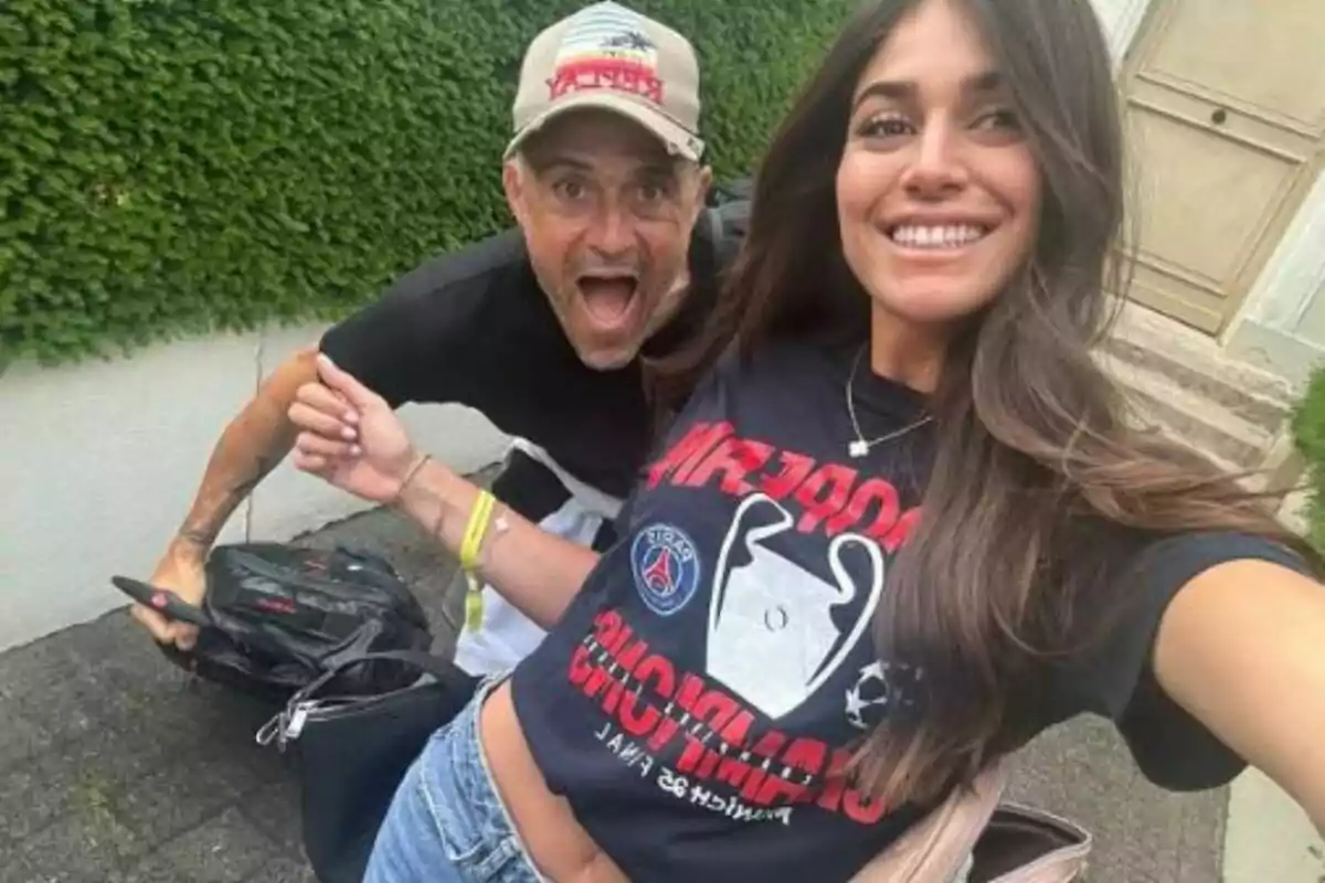A smiling young woman takes a selfie with a man making a funny gesture outdoors in front of a wall covered with plants.