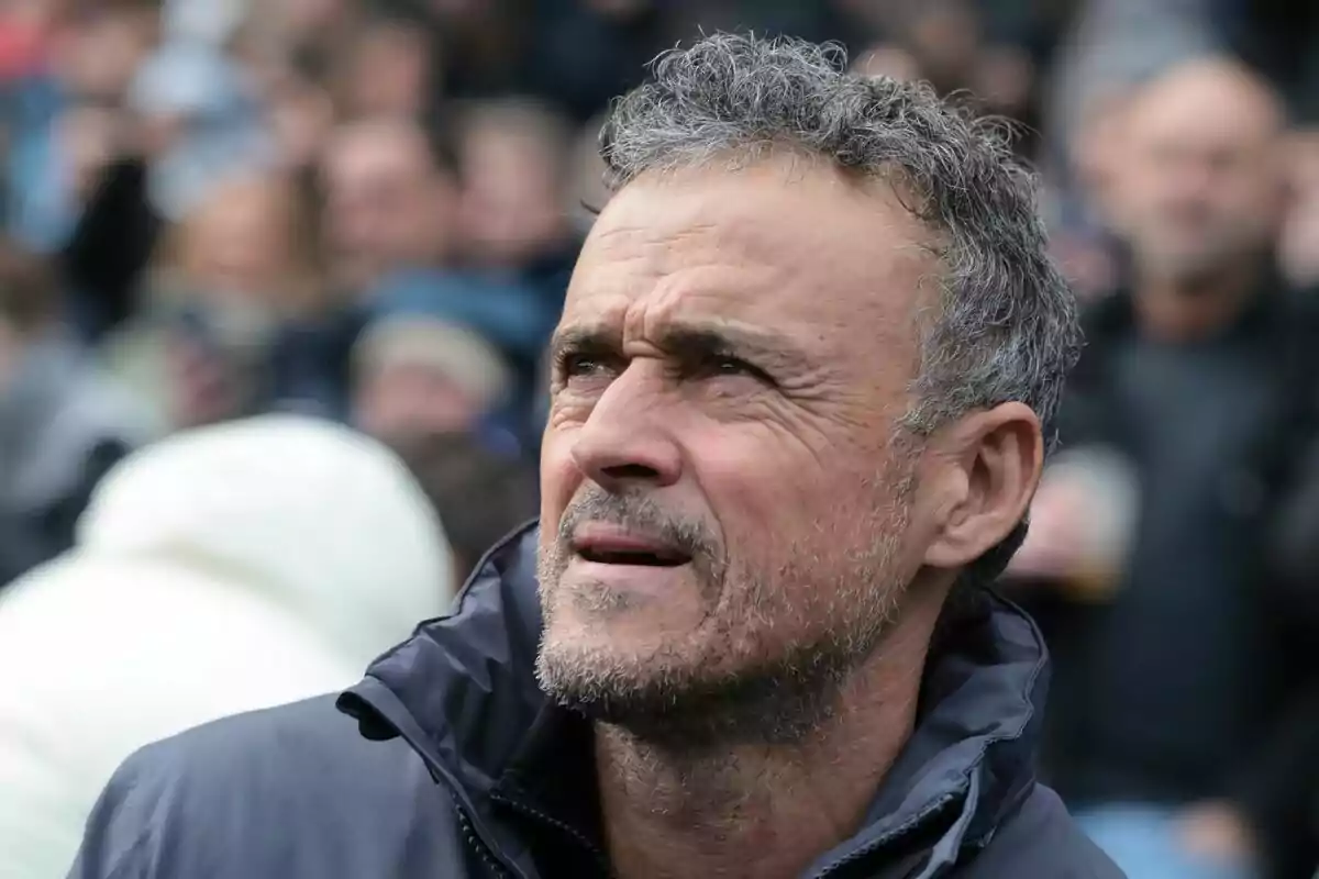 A man in a dark jacket looks up in an outdoor setting with people out of focus in the background.