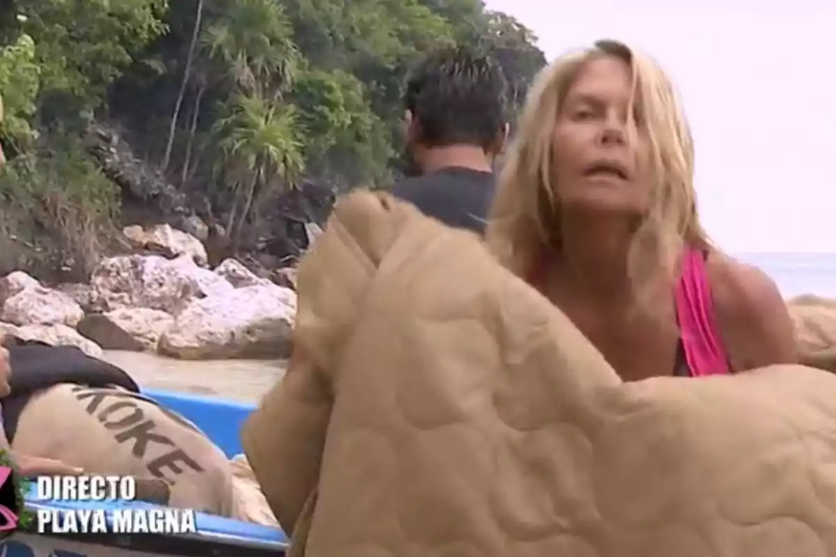 Dona rossa amb samarreta rosa sosté una manta beix a la vora d’una platja amb vegetació i roques al fons mentre una altra persona està d’esquena i es veuen sacs en una barca blava. Dona rossa amb samarreta rosa sosté una manta beix a la vora d’una platja amb vegetació i roques al fons mentre una altra persona està d’esquena i es veuen sacs en una barca blava.