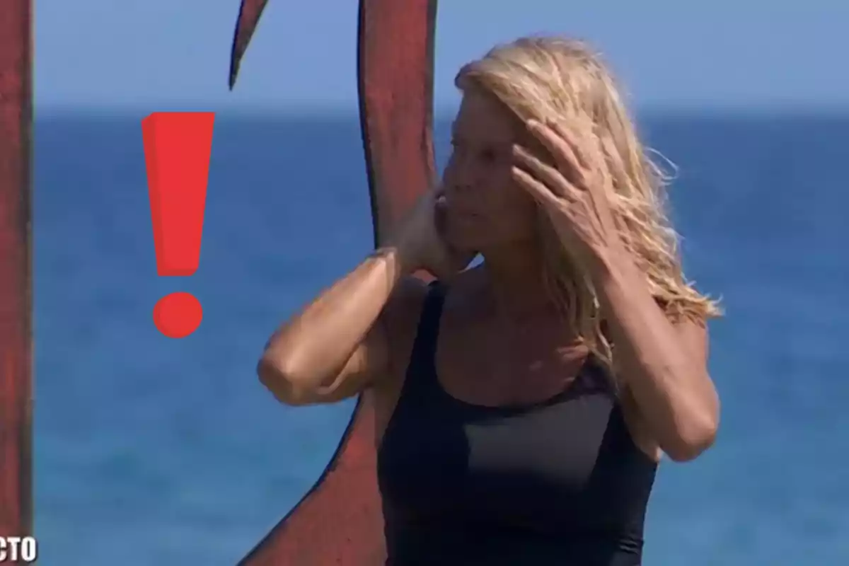 Una persona amb cabell ros i samarreta negra està dempeus al costat del mar, tocant-se el cabell, amb un signe d'exclamació vermell al costat.