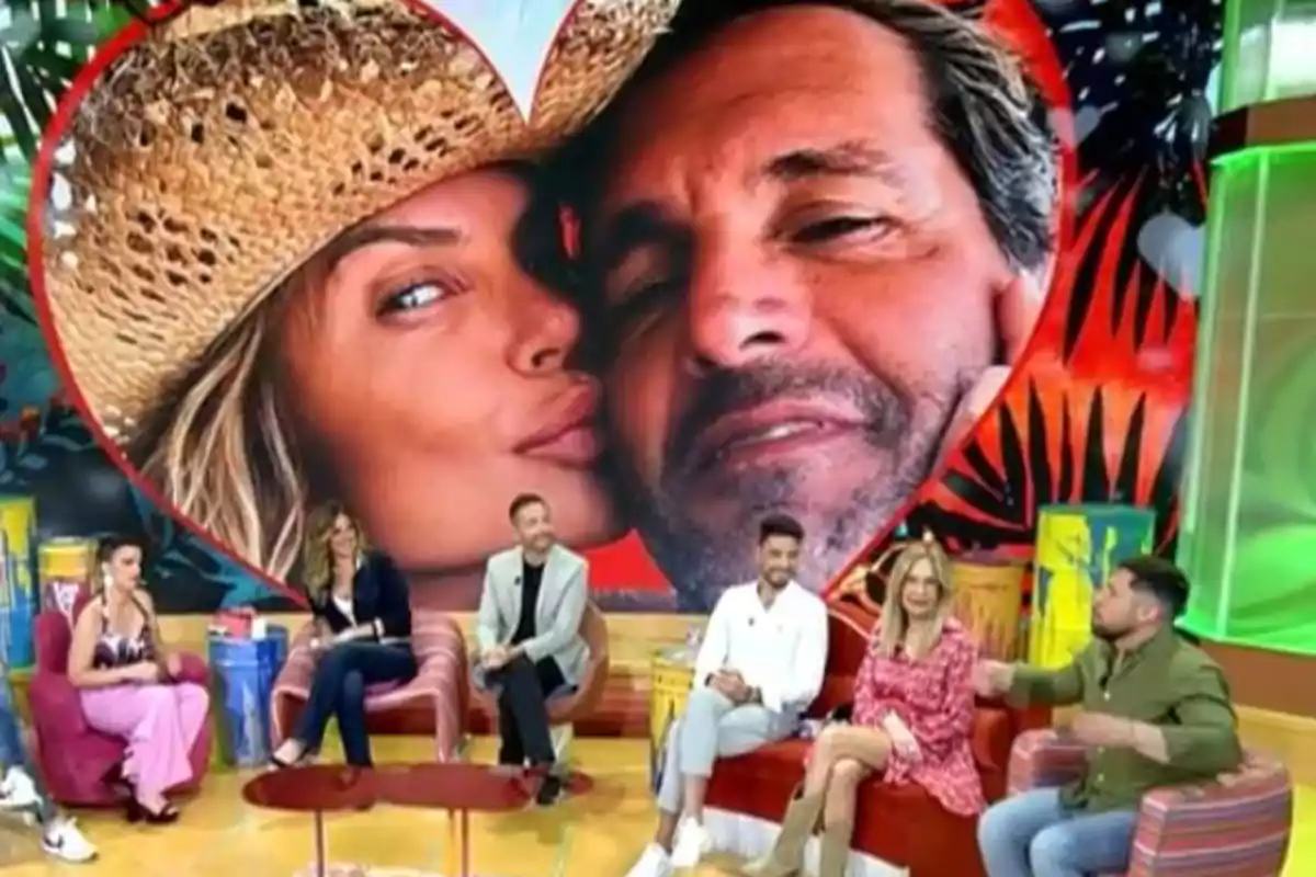 Un grup de persones està assegut en un plató de televisió amb una gran imatge de fons que mostra una parella dins d'un cor. Un grup de persones està assegut en un plató de televisió amb una gran imatge de fons que mostra una parella dins d'un cor.