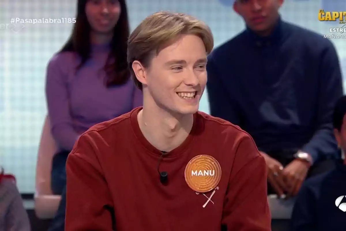 Un jove somrient amb un suèter vermell i una etiqueta que diu “Manu” està assegut en un programa de televisió amb persones al fons.