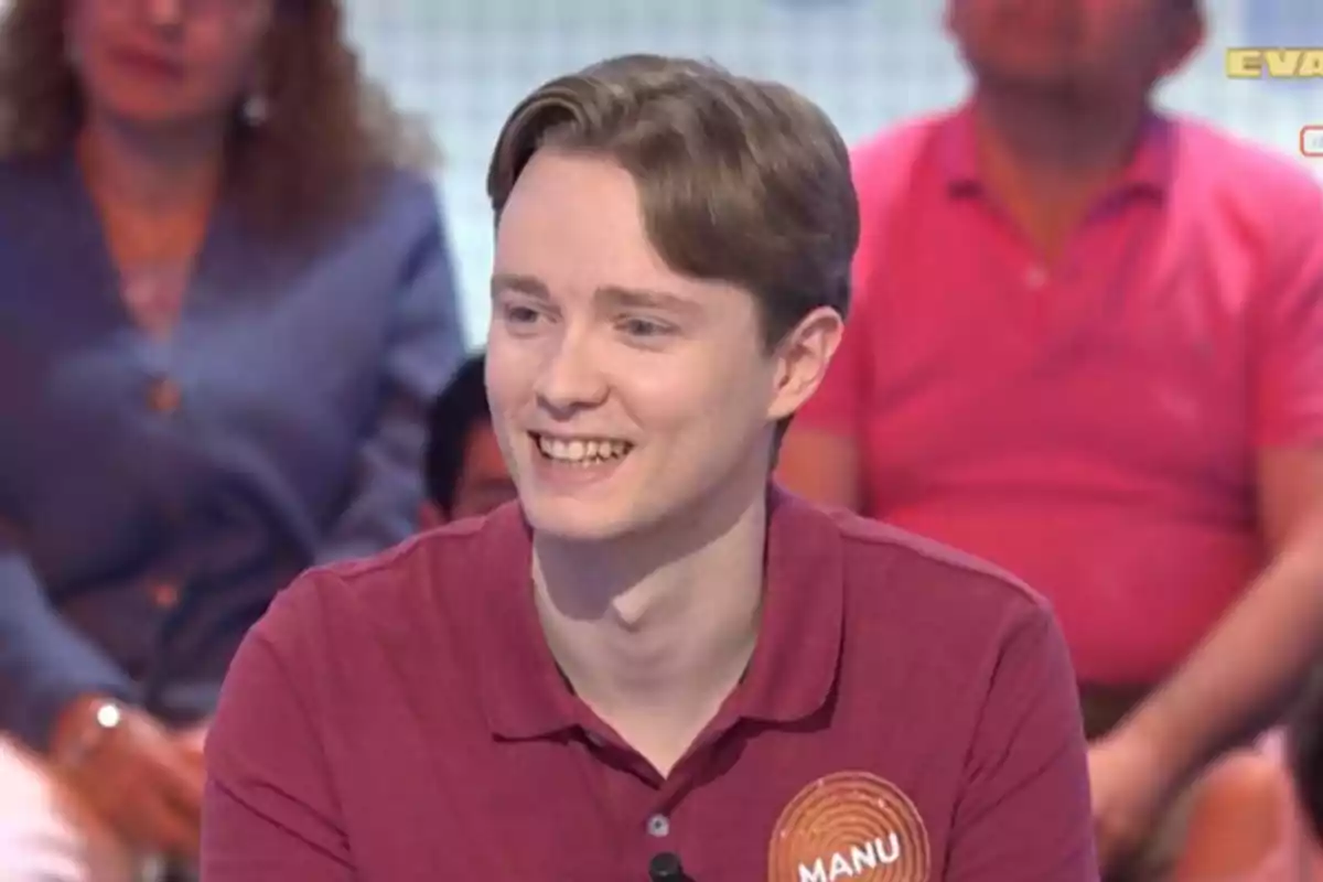 Un jove somrient amb camisa burdeus i una etiqueta que diu Manu participa en un programa de televisió amb públic al fons. Un jove somrient amb camisa burdeus i una etiqueta que diu Manu participa en un programa de televisió amb públic al fons.