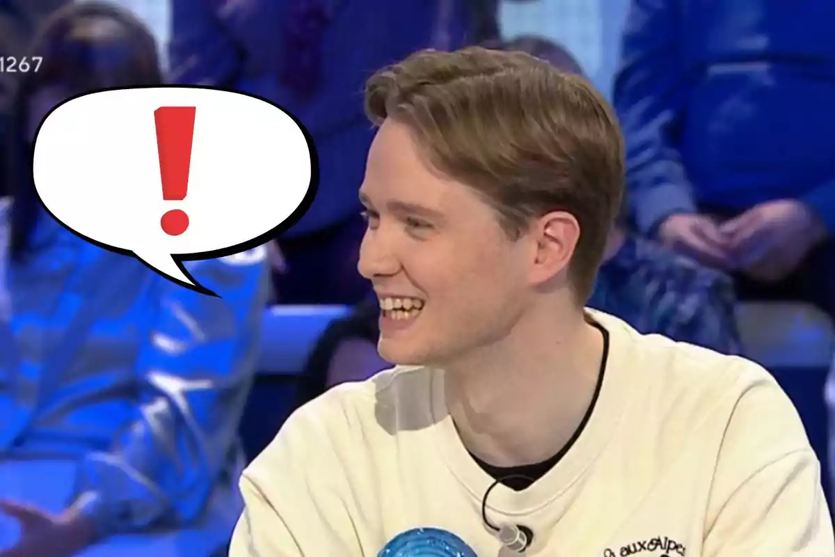 Un home jove somriu mentre està assegut en un programa de televisió, amb un globus de diàleg que conté un signe d'exclamació.