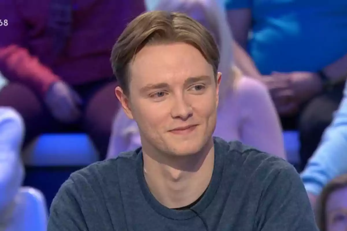 Un home jove amb cabell castany clar i jersei blau somriu mentre està assegut en un programa de televisió amb persones desenfocades al fons. Un home jove amb cabell castany clar i jersei blau somriu mentre està assegut en un programa de televisió amb persones desenfocades al fons.