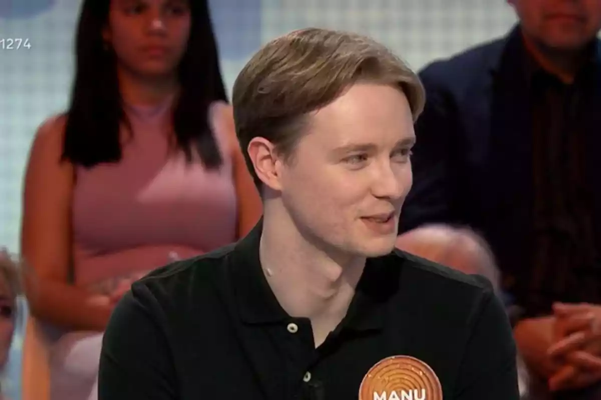Un home jove amb cabell clar i camisa fosca està assegut en un programa de televisió, amb persones al fons. Un home jove amb cabell clar i camisa fosca està assegut en un programa de televisió, amb persones al fons.