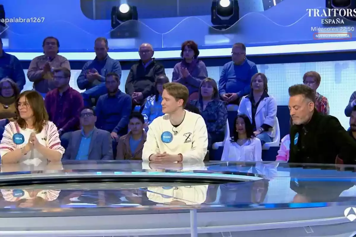 Un grup de persones està assegut en un plató de televisió, amb un públic al fons observant. Un grup de persones està assegut en un plató de televisió, amb un públic al fons observant.