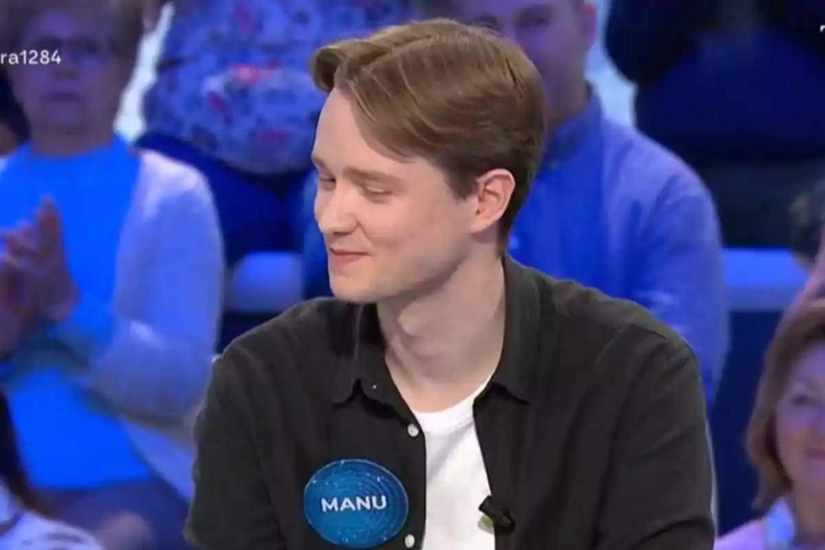 Un jove amb cabell castany clar i camisa negra somriu mentre participa en un programa de televisió, porta una etiqueta blava amb el nom Manu i hi ha públic desenfocat al fons. Un jove amb cabell castany clar i camisa negra somriu mentre participa en un programa de televisió, porta una etiqueta blava amb el nom Manu i hi ha públic desenfocat al fons.