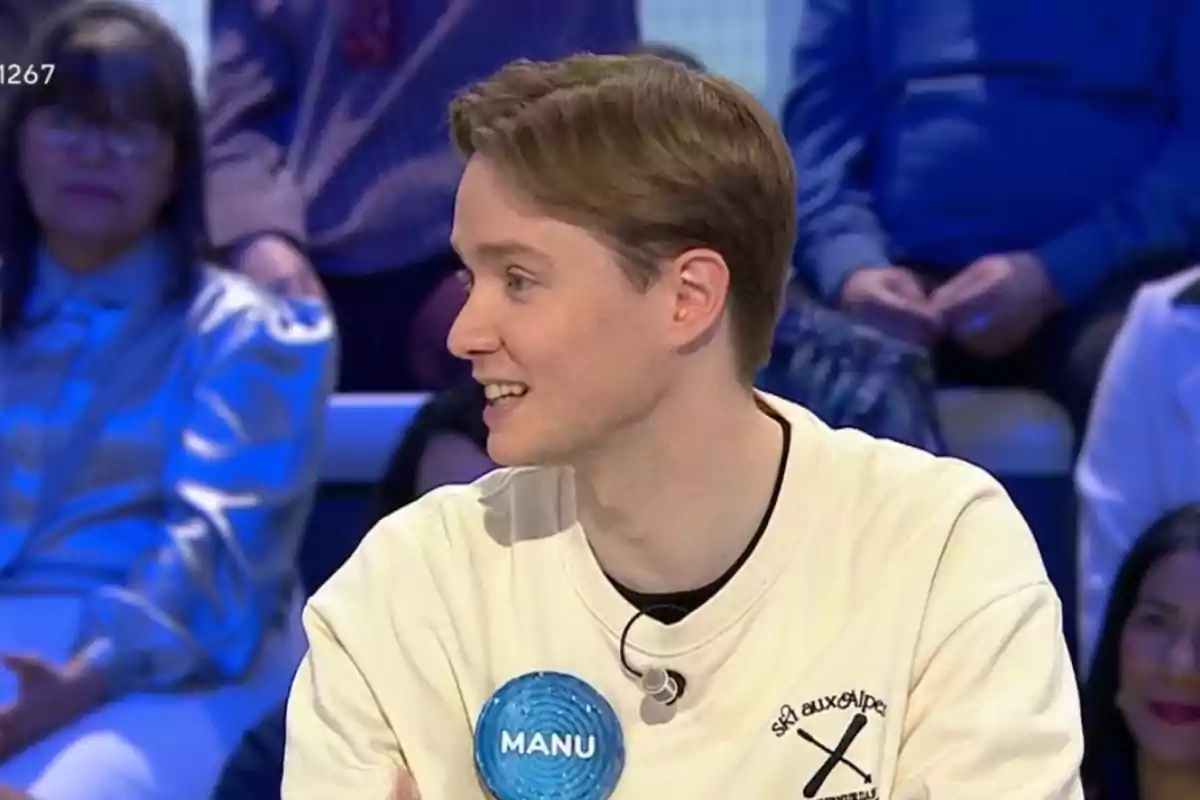 Un home jove amb cabell clar i una samarreta blanca està assegut en un estudi de televisió amb un micròfon a la samarreta i una placa que diu Un home jove amb cabell clar i una samarreta blanca està assegut en un estudi de televisió amb un micròfon a la samarreta i una placa que diu
