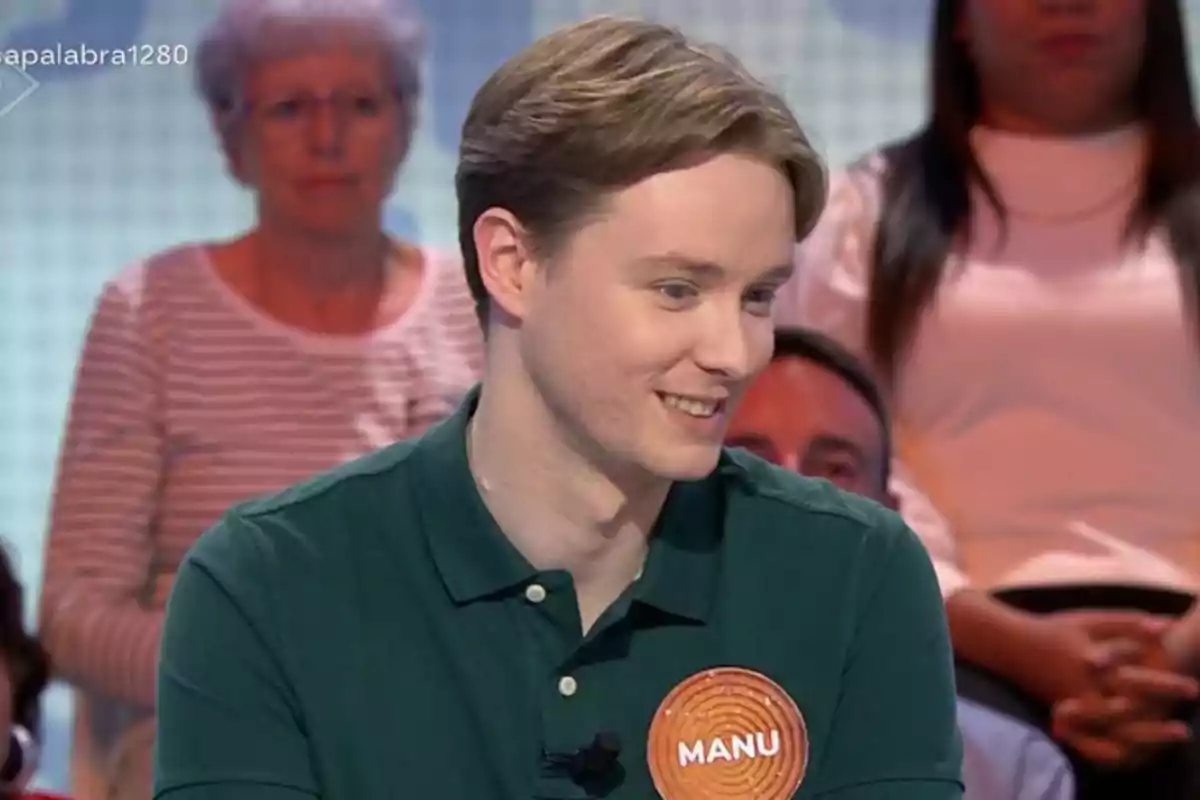 Jove amb camisa verda i una placa que diu Manu participant en un programa de televisió amb públic de fons Jove amb camisa verda i una placa que diu Manu participant en un programa de televisió amb públic de fons