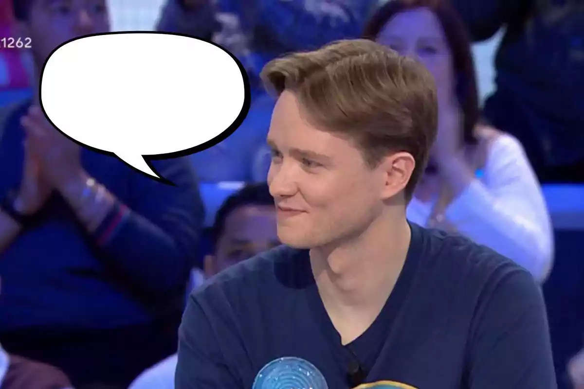 Un home jove amb cabell clar i samarreta fosca està assegut en un estudi de televisió, somrient mentre mira cap a un costat; hi ha un globus de diàleg buit sobre el seu cap i persones desenfocades al fons.