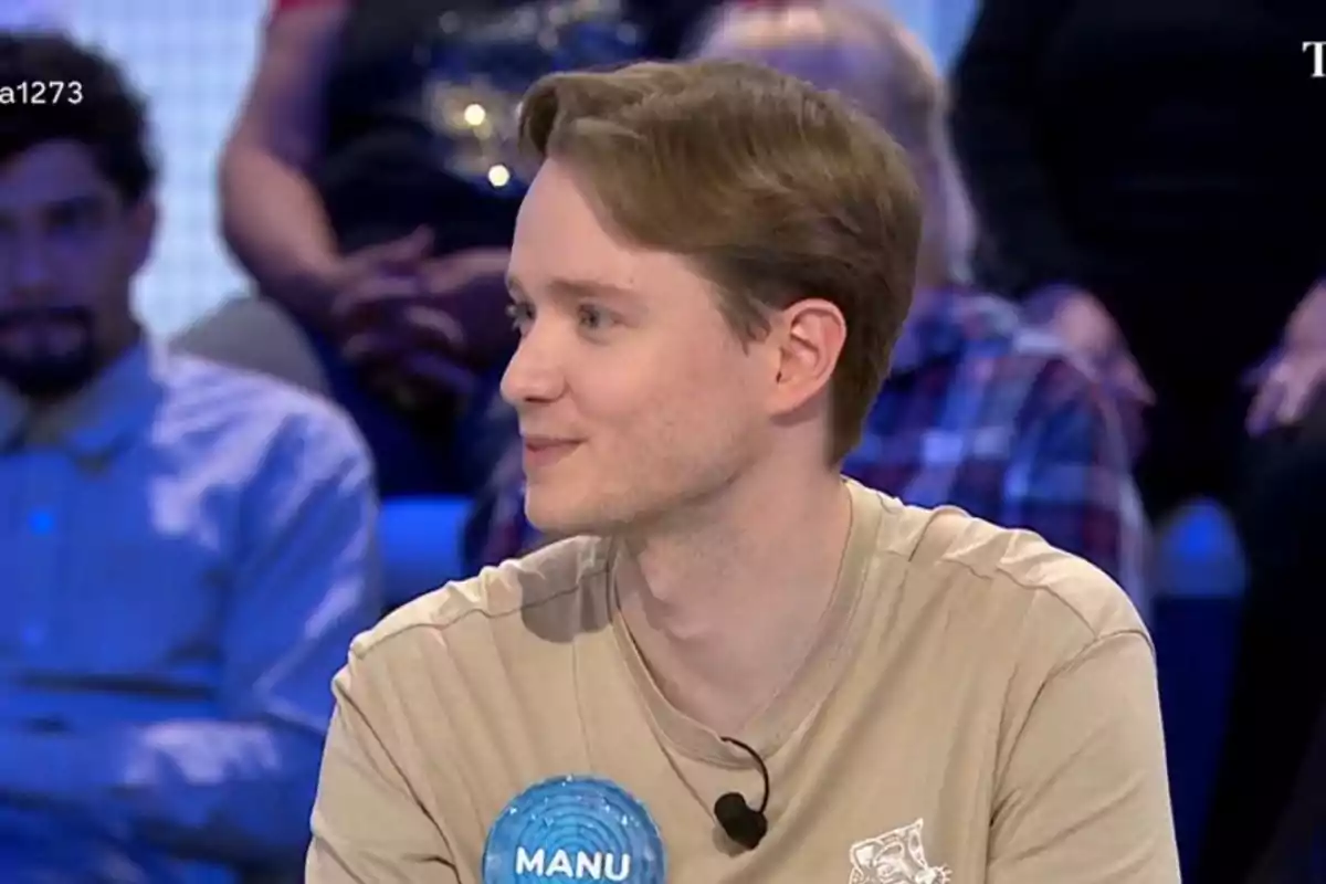 Un home jove amb cabell clar i samarreta beige està assegut en un estudi de televisió amb un micròfon a la solapa i una placa que diu Un home jove amb cabell clar i samarreta beige està assegut en un estudi de televisió amb un micròfon a la solapa i una placa que diu