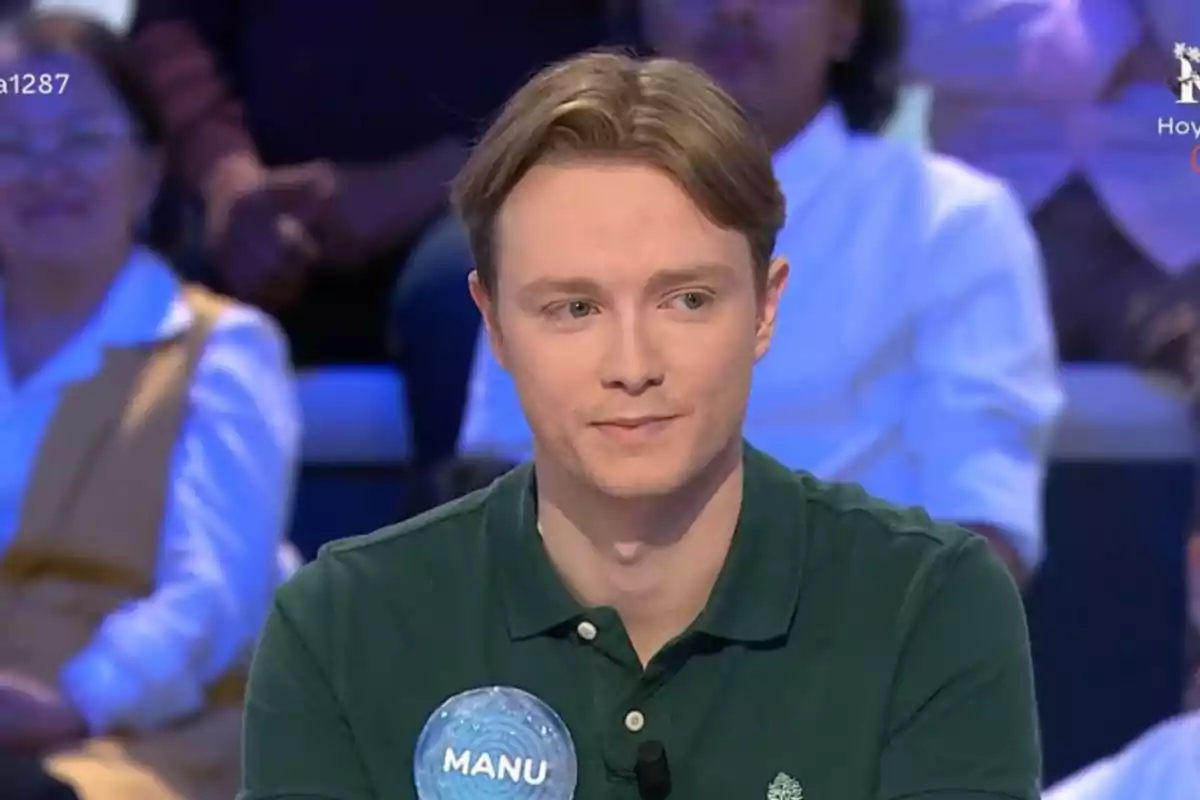 Jove de cabells rossos i camisa verda assegut en un programa de concursos amb públic al fons i una etiqueta que diu Manu al pit Jove de cabells rossos i camisa verda assegut en un programa de concursos amb públic al fons i una etiqueta que diu Manu al pit
